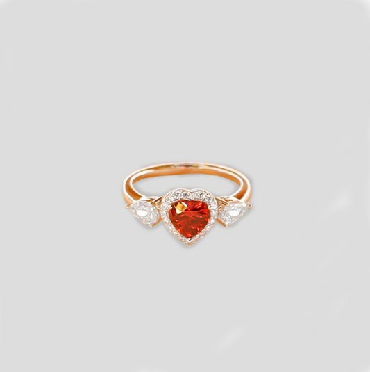 Gold-colored ring featuring a heart-shaped red gemstone at the center, flanked by two teardrop-shaped simulant diamonds.