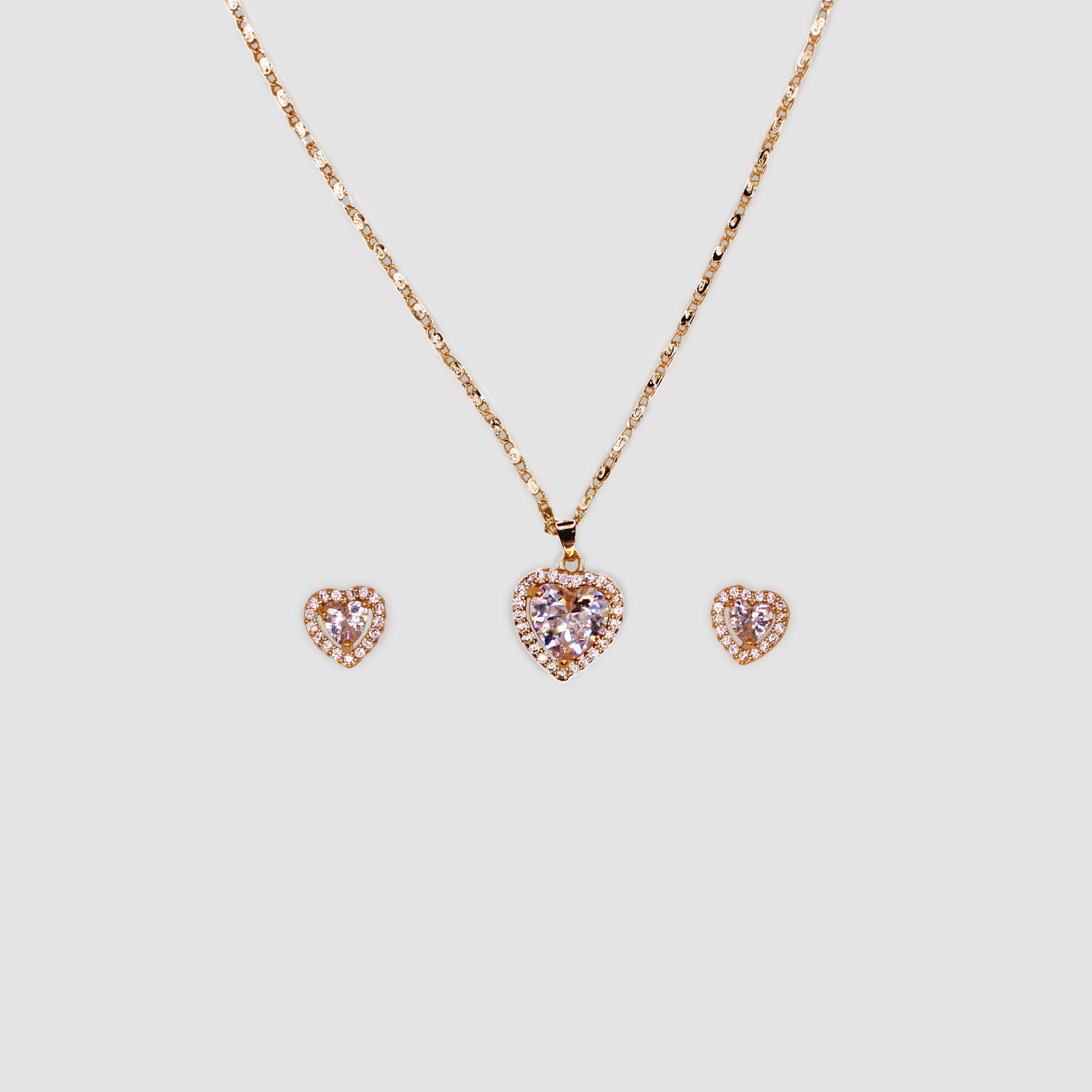 A matching set of gold-colored jewelry featuring a necklace and earrings. The necklace has a heart-shaped pendant with a central clear gemstone surrounded by smaller stones, and the earrings have a similar heart-shaped design with a central gemstone encircled by smaller stones.